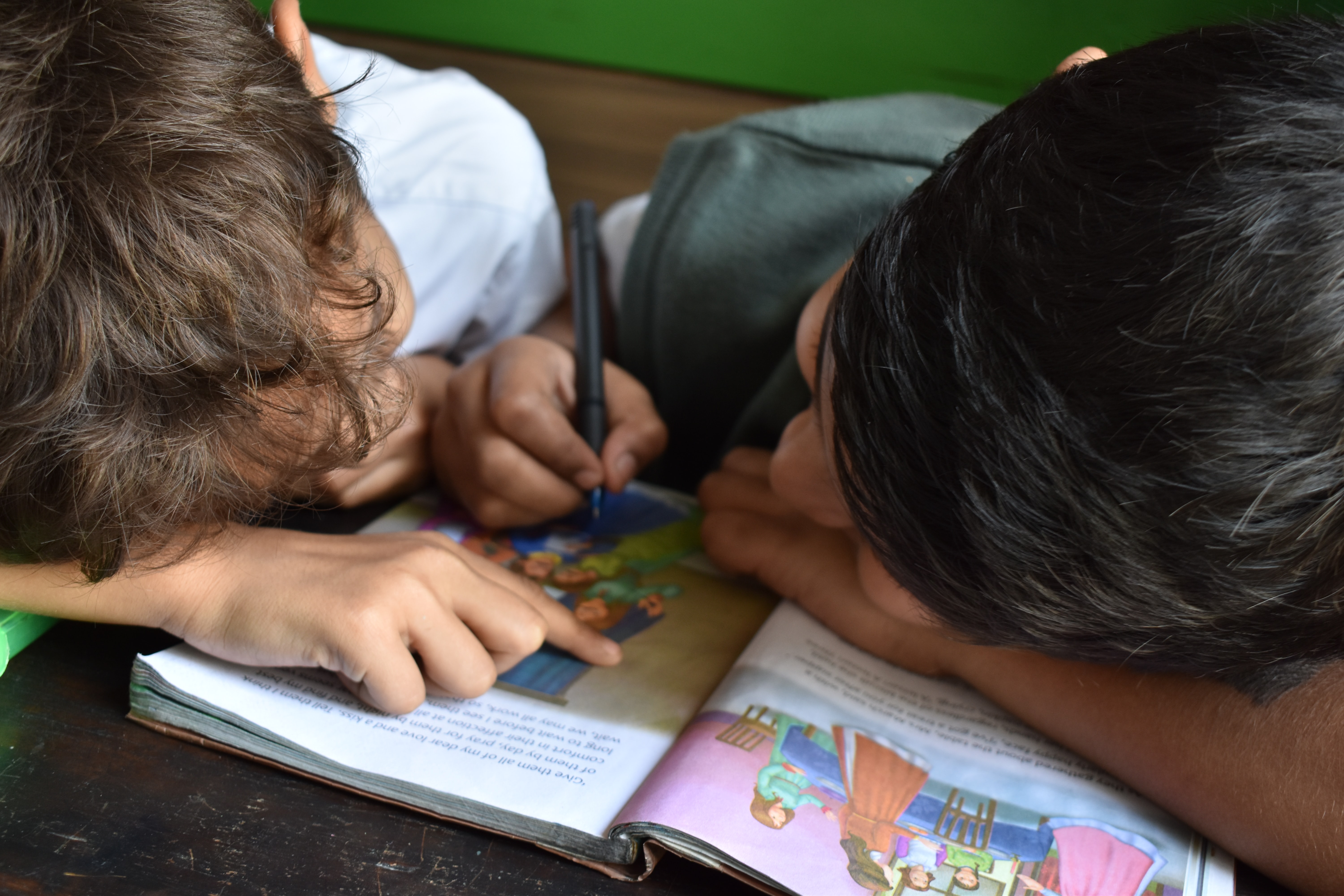 two children leaning over a book together, one using a pen