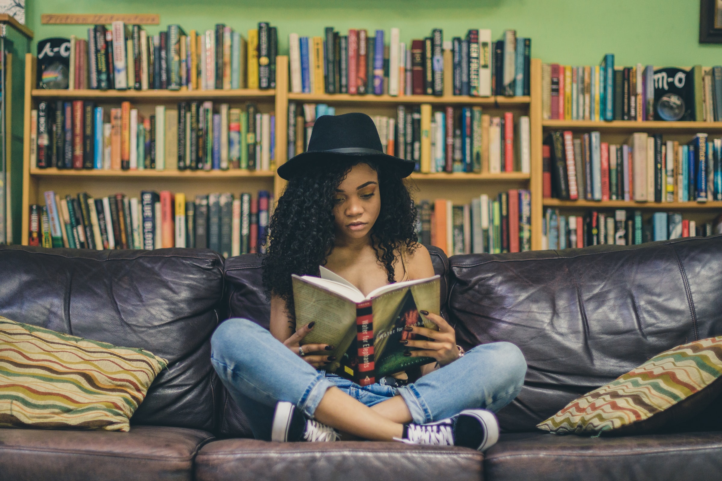 girl sitting on a sofa in front of a bookshelf, wearing a hate and reading a book