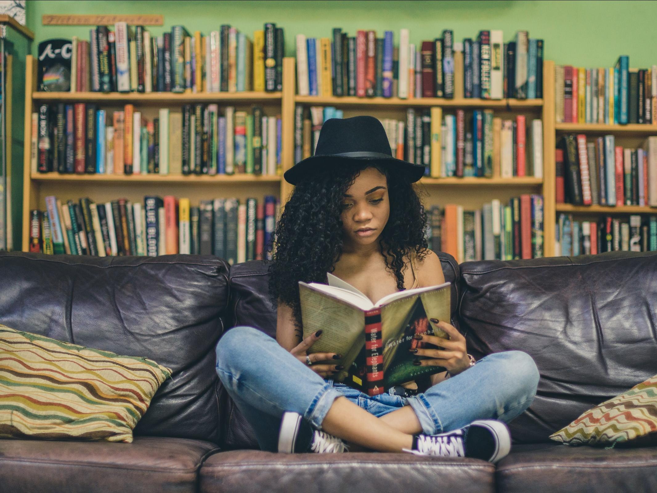 girl sitting on a sofa in front of a bookshelf, wearing a hate and reading a book
